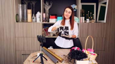 Hediyeler yakın çekimde paketleniyor. Tasarımcı, koruyucu eldivenlerle, akıllı telefonun önünde ipek kurdele ile bir hediye kutusu süslüyor. Online satışlar, webinar. Yüksek kalite fotoğraf