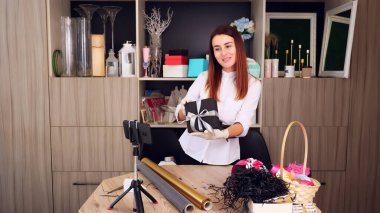 Hediyeler yakın çekimde paketleniyor. Tasarımcı, koruyucu eldivenlerle, akıllı telefonun önünde ipek kurdele ile bir hediye kutusu süslüyor. Online satışlar, webinar. Yüksek kalite fotoğraf