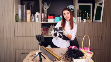 Hediyeler yakın çekimde paketleniyor. Tasarımcı, koruyucu eldivenlerle, akıllı telefonun önünde ipek kurdele ile bir hediye kutusu süslüyor. Online satışlar, webinar. Yüksek kalite fotoğraf