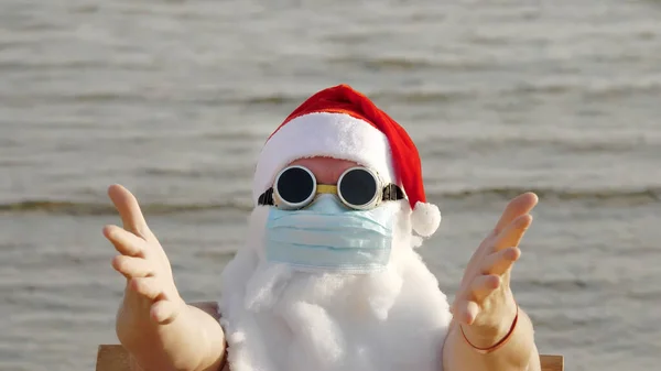面白いサンタ クラス保護マスクだ サンタは サングラスで 手を振って 海のそばのビーチで リラックスした 海岸でのコロナウイルスのパンデミックやロックダウン中のサンタ クラスの休暇 — ストック写真