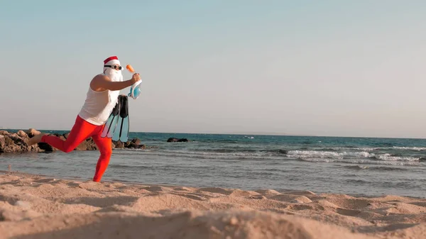 Santa Claus summer vacation. Santa Claus having fun. Funny Santa, in sunglasses, with flippers and snorkeling mask, runs along sea beach. He is in a hurry to swim, snorkel. High quality photo
