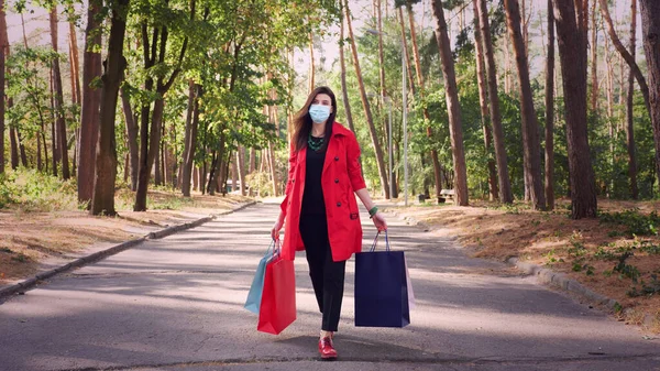 Woman with shopping bags. woman, in protective mask, with colored shopping bags in her hands, walking through city park. shopping and gifts. delivery or donation concept. High quality photo