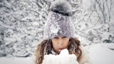 Karlı kış eğlencesi. Kar üfleme. Sıcak kışlık giysiler giymiş mutlu, sevimli, güzel küçük kız, karlı parkta ya da ormanda, kameraya, açık havada ellerinden kar üflüyor. Yüksek kalite fotoğraf