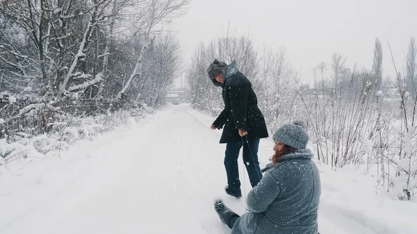 family sledding in winter. outdoor winter activity. Happy, laughing, playful married couple is enjoying of sledging on snowy road, in forest, during snowfall. They having fun, spending time together