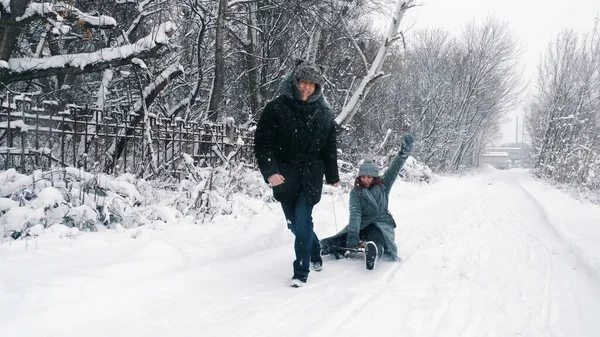 family sledding in winter. outdoor winter activity. Happy, laughing, playful married couple is enjoying of sledging on snowy road, in forest, during snowfall. They having fun, spending time together