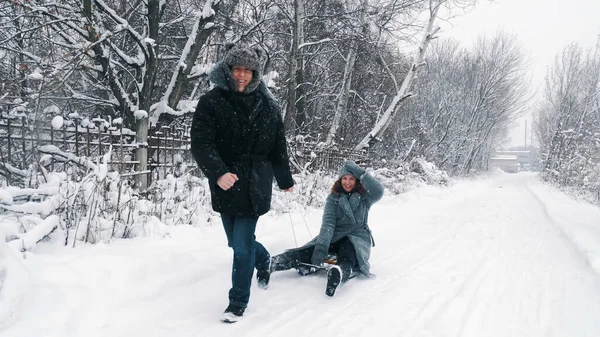 family sledding in winter. outdoor winter activity. Happy, laughing, playful married couple is enjoying of sledging on snowy road, in forest, during snowfall. They having fun, spending time together