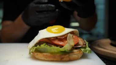 Hamburger. Yemek pişirmek. fast food. Aşçı yumurta burger yapıyor. Sosu, eti ve sebzesi olan sulu bir burger, üstünde kızarmış yumurta ve çıtır ekmek.