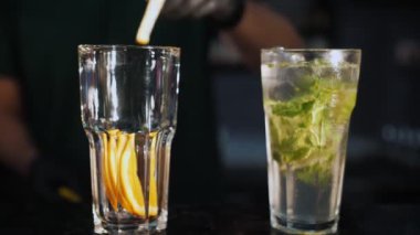 Kokteyl. Barmen kokteyl yapar. Yakın plan. Barmen nane yapraklarını ve portakal dilimlerini bir bardakta ezer. Siyah arka planda iki kokteyl.