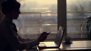 Genç odaklı iş kadını gözlük, karanlık siluet bir şey onun mobil olarak yazarak, sonraki tablo üstünde laptop. gün batımında, ışınları içinde bir büyük ofis pencere arka planı