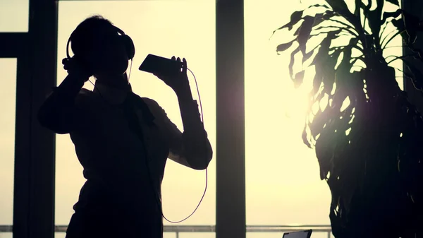 dark silhouette of business woman, dances with headphones and mobile in her hand, listen music, against the background of a large office window, at sunset, in the rays of light. office break. High