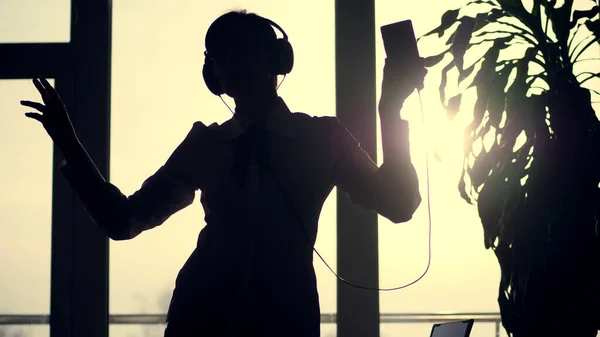 dark silhouette of business woman, dances with headphones and mobile in her hand, listen music, against the background of a large office window, at sunset, in the rays of light. office break. High