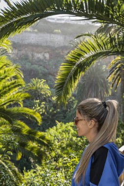 Beyaz kadın ince mavi ceketi ve palmiye ağaçları arasında uzun sarı saçları var.