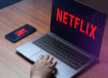 Genç bir kadın dizüstü bilgisayardan Netflix izliyor. Dizüstü bilgisayarda Netflix logosu ve akıllı telefon ekranları. Dizüstü bilgisayarlı ahşap masa ve Netflix 'te akıllı telefon yayını.