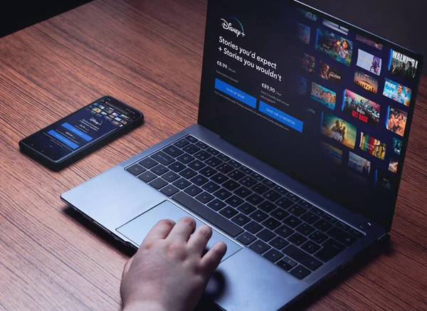 Disney + dizüstü bilgisayarda ve akıllı telefon ekranlarında. Dizüstü bilgisayarlı ahşap masa ve Disney + 'dan akıllı telefon yayını. Disney izlemek için dizüstü bilgisayar kullanan genç bir kadın. +.