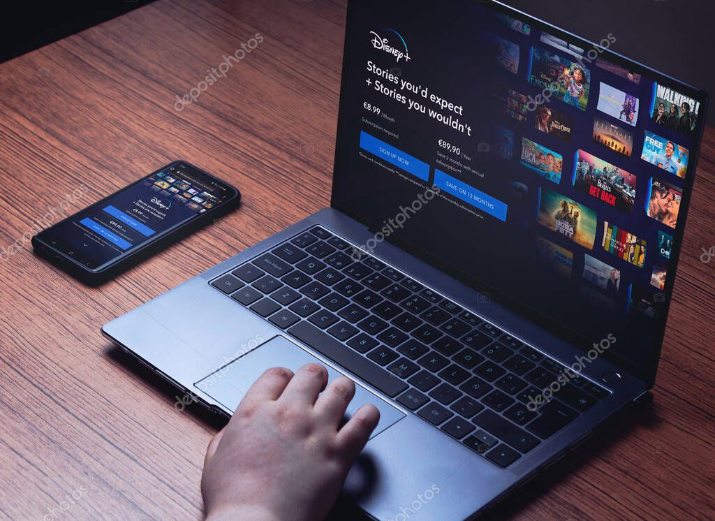Disney+ on laptop and smartphone screens. Wood desk with laptop and smartphone streaming Disney+. Young woman using laptop to watch Disney +.