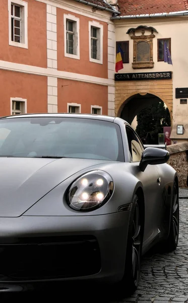 Trunted spor araba, şehirde küçük bir caddeye park edilmiş, farlar açık. Arka planda renkli binalar görünür..