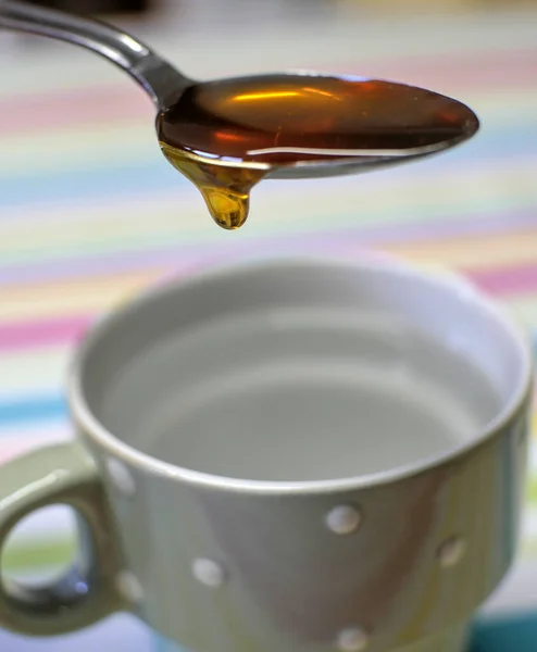 Tea spoon filled with liquid honey, a drop of which will fall into the mug of water below.