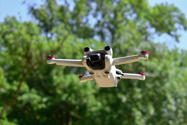 Small white mini drone flying up in front of forest, shallow depth of field