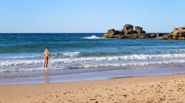 Sarışın kız sörf yapan erkek arkadaşını izliyor, okyanus dalgaları kayalara çarpıyor, Praia do Castelo Sahili, Algarve, Güney Portekiz, Avrupa