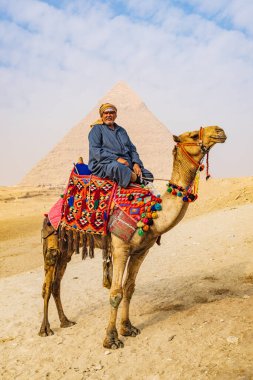Giza, Kahire, Mısır. 18 Şubat 2022. Battaniyesi sökülmüş bir devenin üzerindeki adam ve Giza 'daki Büyük Piramit kompleksinde yular..