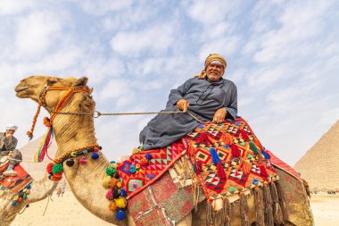 Giza, Kahire, Mısır. 18 Şubat 2022. Battaniyesi sökülmüş bir deveye yaslanmış bir adam ve Giza 'daki Büyük Piramit kompleksinde yular..