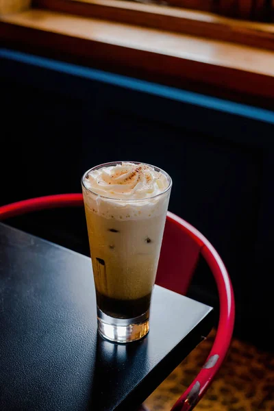 A glass of iced hojicha chocolate latte in a corner of a table with dark and blurred background. Behind it, there is a red chair.
