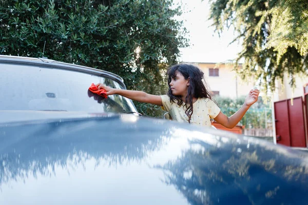 Girl with long brown hair is washing blue vintage car in the garden with orange cloth