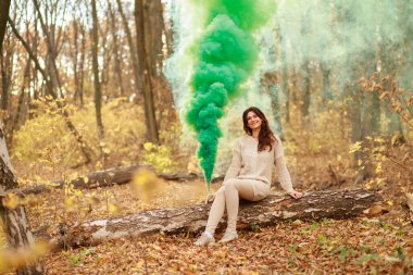 Genç ve güzel bir kadın sonbahar ormanında bir ağaç dalında oturuyor ve elinde yeşil dumanlı bir maytap tutuyor..