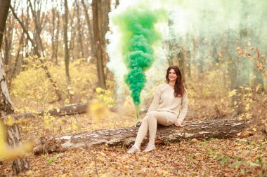 Genç ve güzel bir kadın sonbahar ormanında bir ağaç dalında oturuyor ve elinde yeşil dumanlı bir maytap tutuyor..