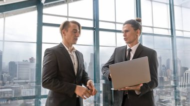İki yönetici, şehir manzaralı bir terasta yeni proje hakkında gayri resmi bir konuşma yaparken bilgisayar defteri kullanıyor. İş dünyası liderleri yeşil bir binada rahatlatıcı atmosfer altında fikir alışverişinde bulunurlar..