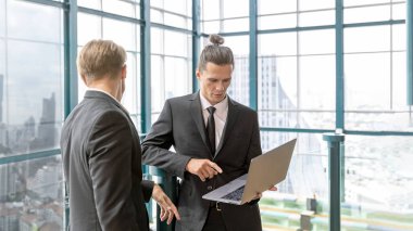 İki yönetici, şehir manzaralı bir terasta yeni proje hakkında gayri resmi bir konuşma yaparken bilgisayar defteri kullanıyor. İş dünyası liderleri yeşil bir binada rahatlatıcı atmosfer altında fikir alışverişinde bulunurlar..