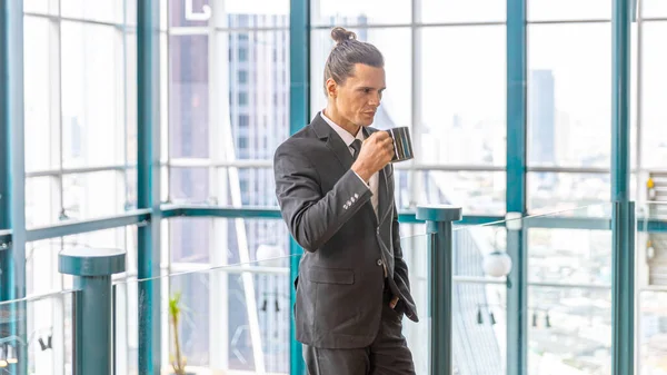 Young SMART CEO with topknot hairstyle takes a cup of coffee at a rooftop terrace with skyscrapers background in an urban or city area. A businessman stands next to window with cityscape background