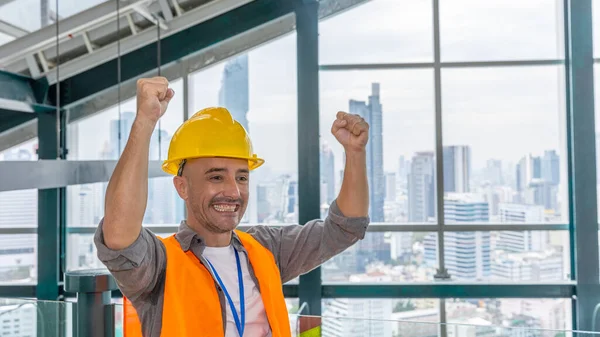 A construction or civil engineer with safety suit expresses his motivation after his project meets target. Foreman in safety uniform stands next to window with cityscape and skyscrapers as background. Young man with construction helmet is very happy