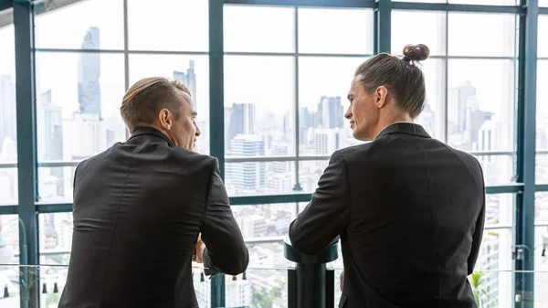 Two managers have an informal conversation about new project at a terrace with cityscape background. Business leaders exchange ideas under relax atmosphere in a green building. Skylines background