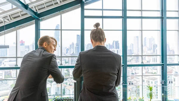 Two managers have an informal conversation about new project at a terrace with cityscape background. Business leaders exchange ideas under relax atmosphere in a green building. Skylines background