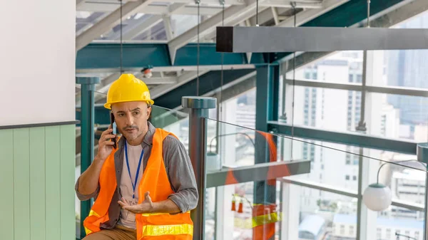 A construction or civil engineer with safety suit have a call with his customer next to window with cityscape and skyscrapers as background.