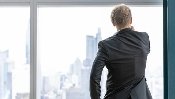 Young CEO stands next to the window showing background of cityscape with high skyscrapers in a city. Confident male business officer gets inspiration from city landscape for future plan