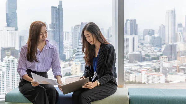 Two female office workers using a laptop to discuss for a new business project in an office with cityscape background. Informal discussion between two young staff with urban landscape background