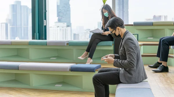 Young business man in generation z with self confidence wearing face mask and using digital device or smartphone to connect with other colleagues at a green corner with skyscraper background