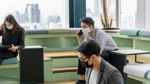 Young business man in generation z with self confidence wearing face mask and using digital device or smartphone to connect with other colleagues at a green corner with skyscraper background