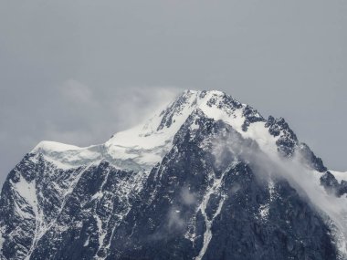 Atmosferik alp manzarası, karla kaplı dağın tepesi, kar altında gri gökyüzü. Güzel, sivri tepeli, karlı ve alçak bulutlu, yüksek karlı dağ duvarlı muhteşem bir manzara..