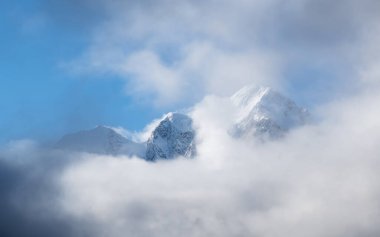 Yumuşak odaklanma. Yüksek karlı dağlı, alçak bulutların üstünde zirvesi ve buzulu olan panoramik alp güneşli manzara. Büyük kar, gün ışığıyla kaplı dağları kapladı. Beyaz karlı, sivri uçlu güneş ışığı.