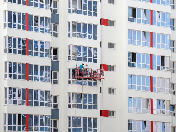 Russia. Barnaul. July 2021. Winch with workers on the facade of a new building under construction. Fragment of a wall with windows of a multi-storey apartment building.