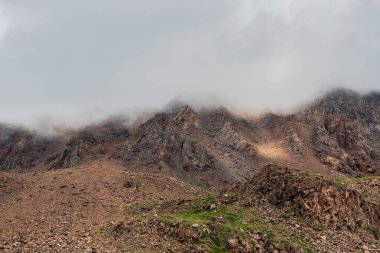 Dağın tepesi alçak bulutların altında. Yüksek dağları olan doğal bir arka plan, kayalarla kaplı dik bir dağ yamacı..