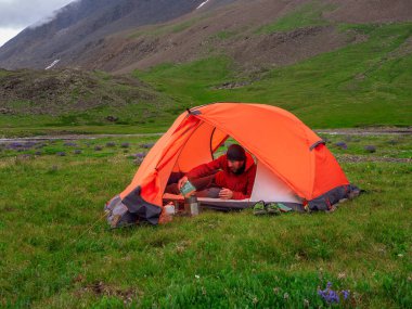 Solo trekking. Camping on a summer green rocky high-altitude plateau. Man in a orange tent is preparing food. Peace and relaxation in nature. 