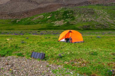 Solar battery at the campsite. Solo hiking in the picturesque summer mountains. Camping on a summer green rocky high-altitude plateau.