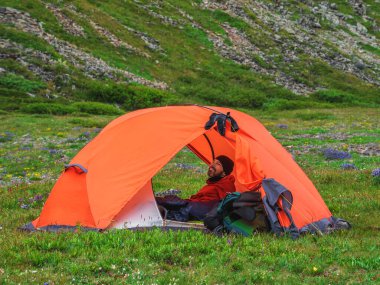 Portrait of a smiling bearded tourist sitting in a tent. Solo trekking. Camping on a summer green rocky high-altitude plateau. Peace and relaxation in nature. 