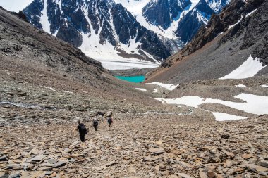 Group of climbers descends a loose slope from the mountain to a distant blue alpine lake. Travel lifestyle, hiking hard track, adventure concept in summer vacation.