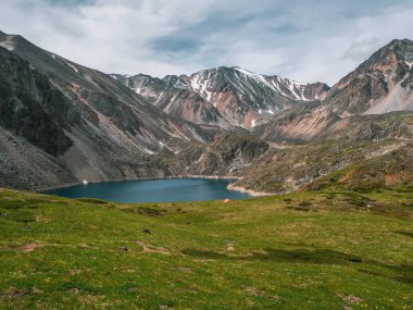 Atmospheric mountain landscape with a small orange tent near an alpine lake. Stunning green landscapes in a high-altitude valley with a clear mountain lake overlooking the snowy mountains.
