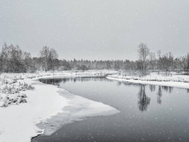 Snowfall on the river. Winter landscape with small river in wood on background gray snowy sky.